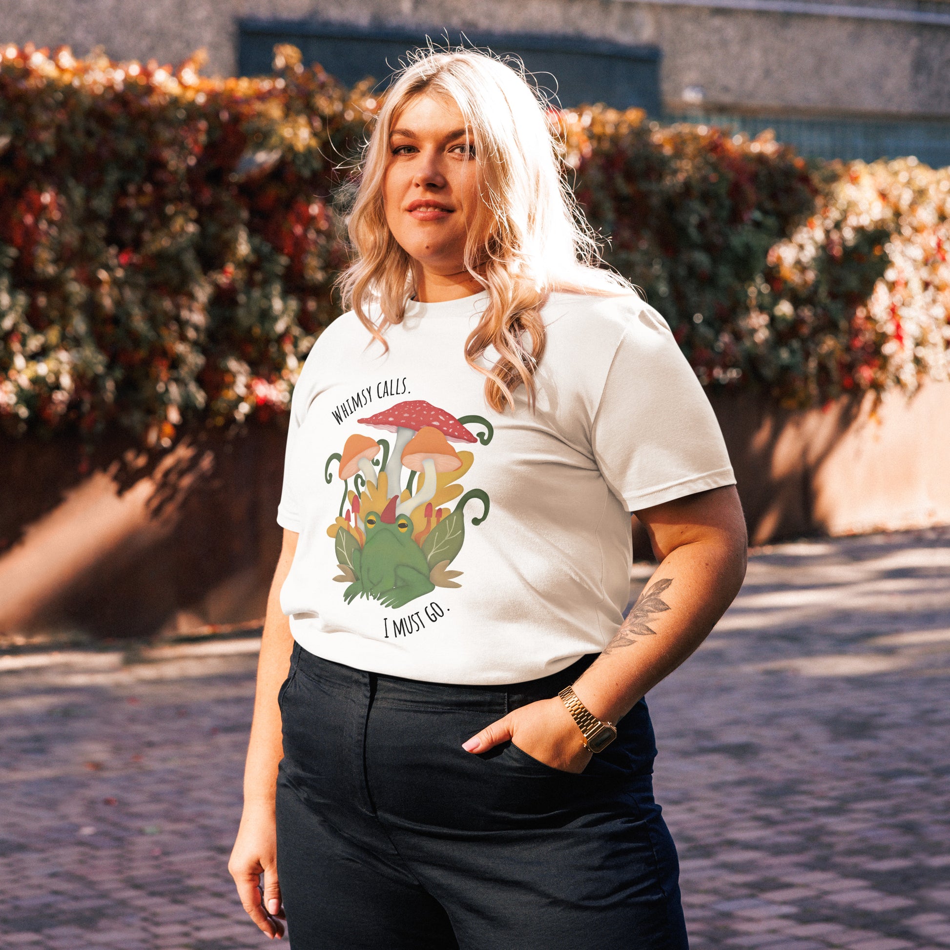 Person wearing a white graphic t-shirt outside with an illustrated design of a frog amongst mushrooms and leaves. Words on the t-shirt read whimsy calls I must go