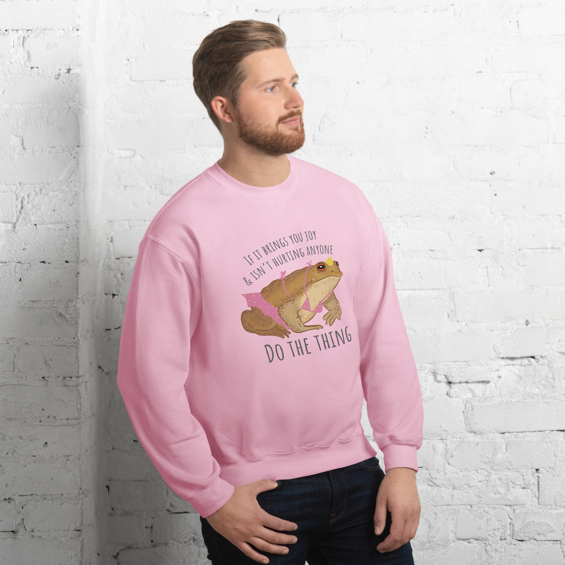 Man wearing a pink sweatshirt with a graphic of a toad and text against a white brick wall.